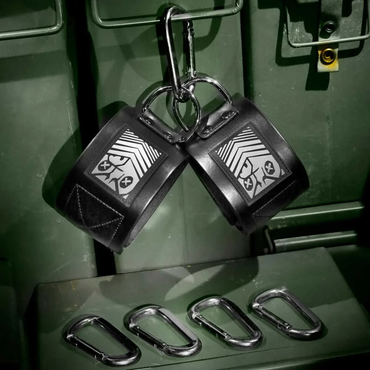 Pair of Neo neoprene ankle cuffs hanging from carabiners on a green ammo crate, 4 clips below, Fort Troff gay gear at Boyzshop