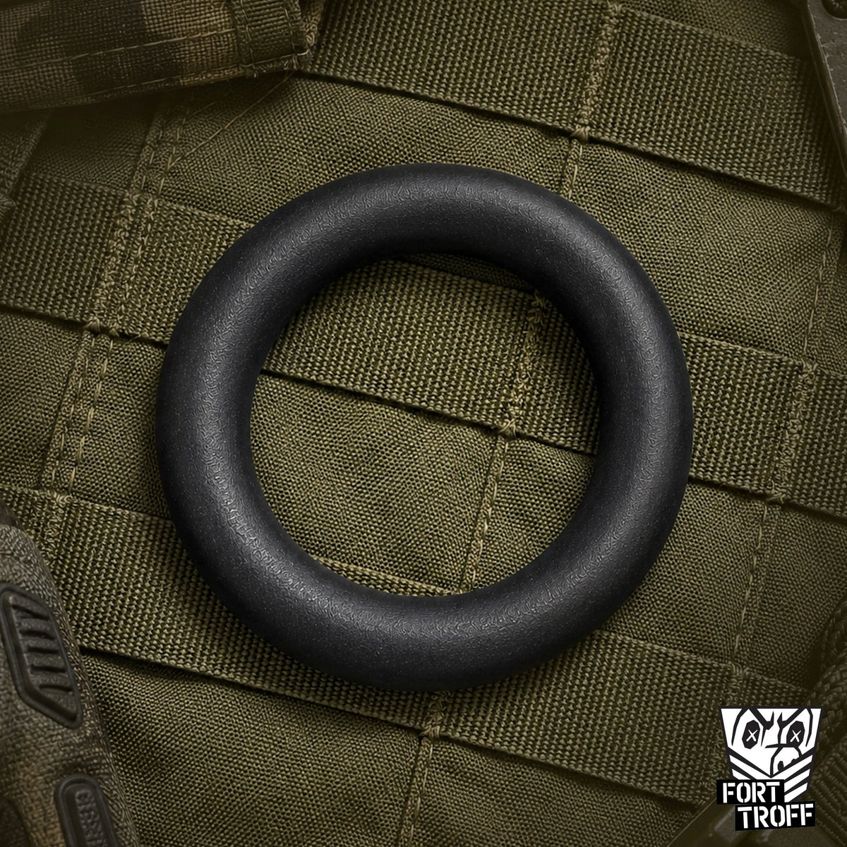 X-Wrap black silicone cock ring lying flat on olive tactical webbing, Fort Troff gay gear at Boyzshop