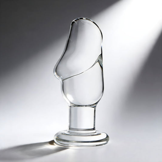 clear glass butt plug with sculpted cock head shape and flared round base displayed on gray background with dramatic lighting
