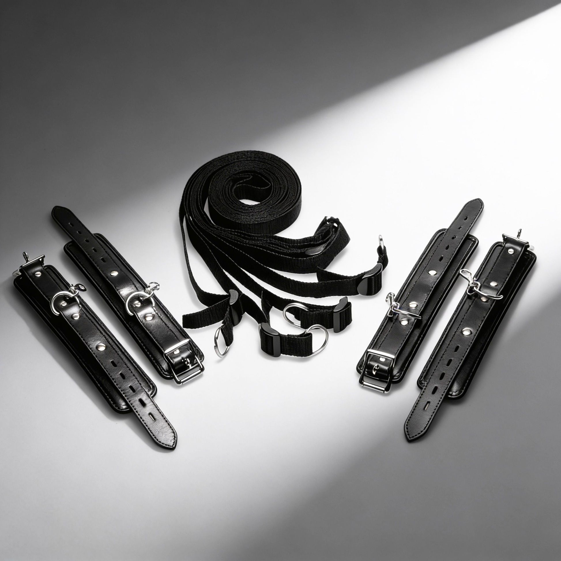 black under-bed restraint kit with adjustable straps, padded cuffs, buckles, and silver clip hardware displayed on white background