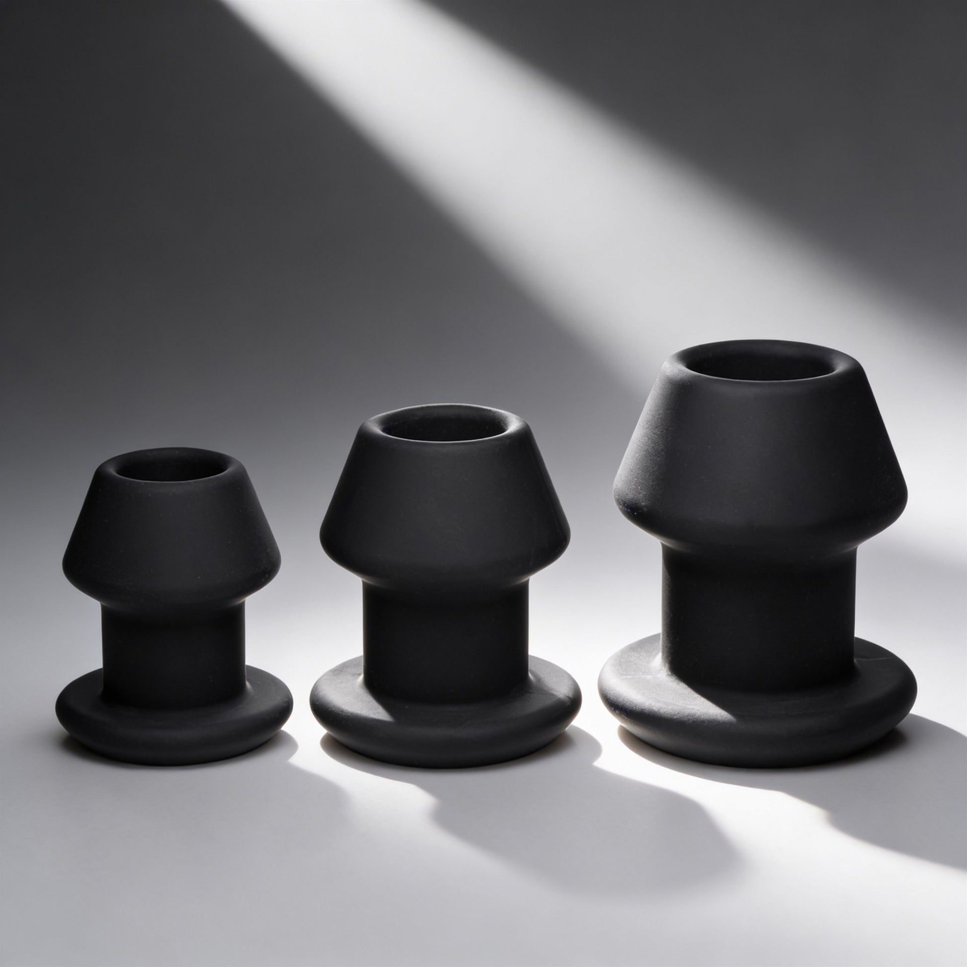 three black hollow silicone anal dilator plugs in graduated sizes with flared bases displayed on gray background under dramatic lighting