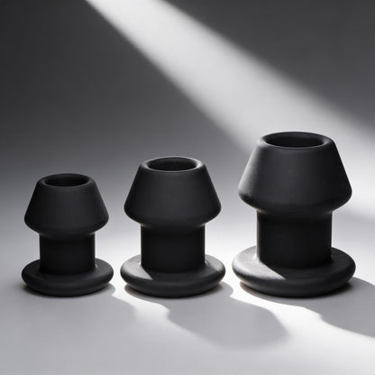 three black hollow silicone anal dilator plugs in graduated sizes with flared bases displayed on gray background under dramatic lighting