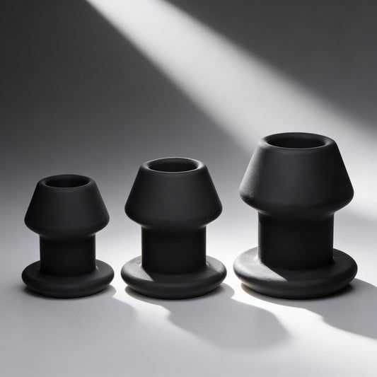 three black hollow silicone anal dilator plugs in graduated sizes with flared bases displayed on gray background under dramatic lighting