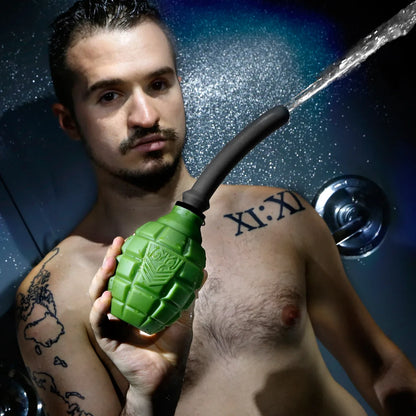 Tattooed model in a shower holding the green Grenade anal douche bulb as water sprays out, Fort Troff gay gear at Boyzshop