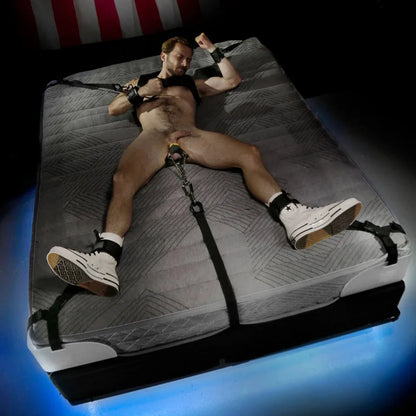 Nude model spread on a mattress, wrists and ankles strapped to the corners, ball strap pulled tight, Fort Troff gay gear at Boyzshop