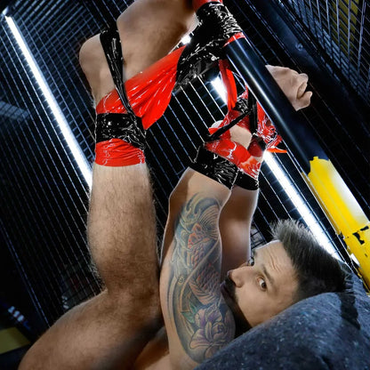 Tattooed model upside down, legs plus arms bound overhead in red bondage tape, Fort Troff gay gear at Boyzshop