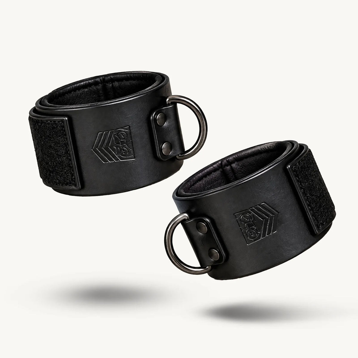 Pair of black leather ankle cuffs closed into loops on white, dark D-rings facing out, Fort Troff gay gear at Boyzshop