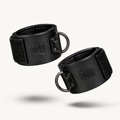 Pair of black leather ankle cuffs closed into loops on white, dark D-rings facing out, Fort Troff gay gear at Boyzshop