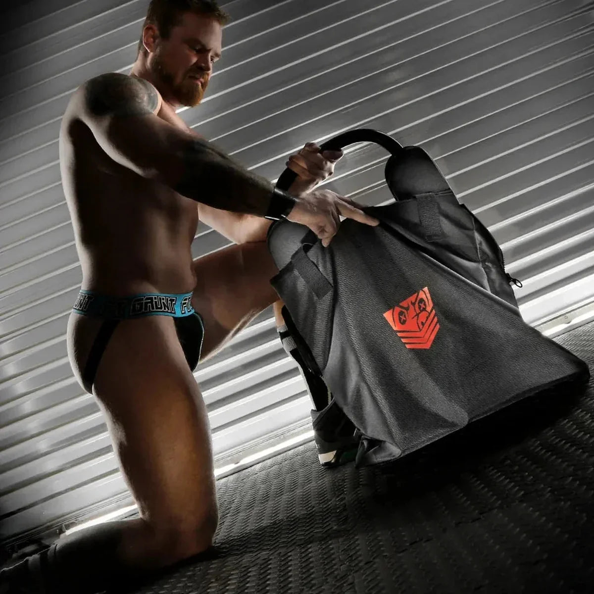 Model kneeling to zip a rim seat into the Rimmer Bag, steel shutter backdrop, Fort Troff gay gear at Boyzshop
