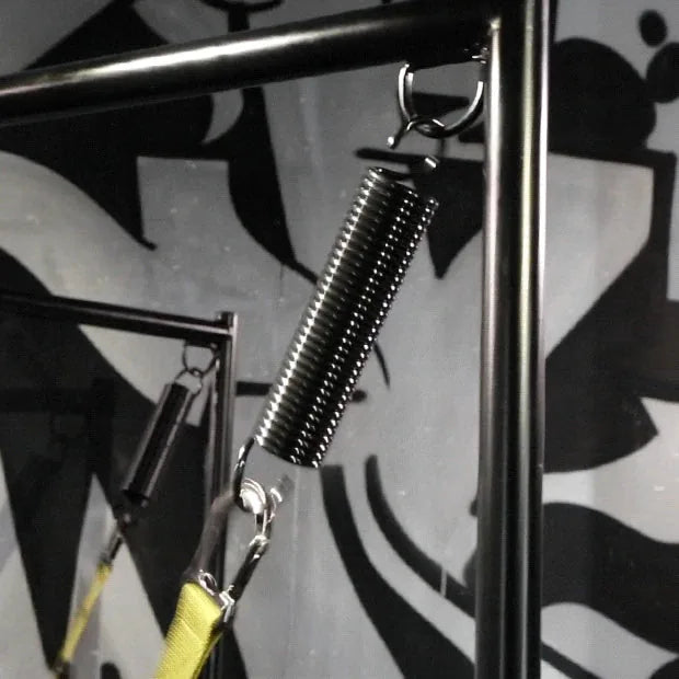 Sling spring close-up showing the coil plus hook on a black sling stand, graffiti wall, Fort Troff gay gear at Boyzshop