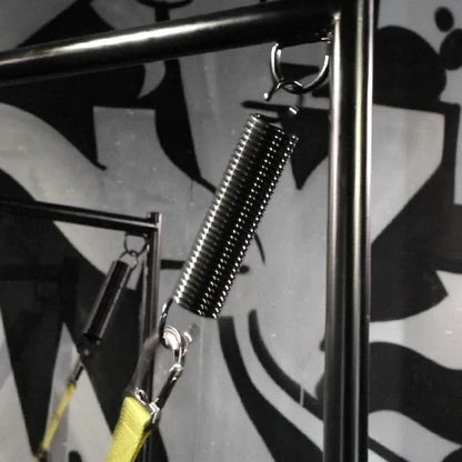 Sling spring close-up showing the coil plus hook on a black sling stand, graffiti wall, Fort Troff gay gear at Boyzshop