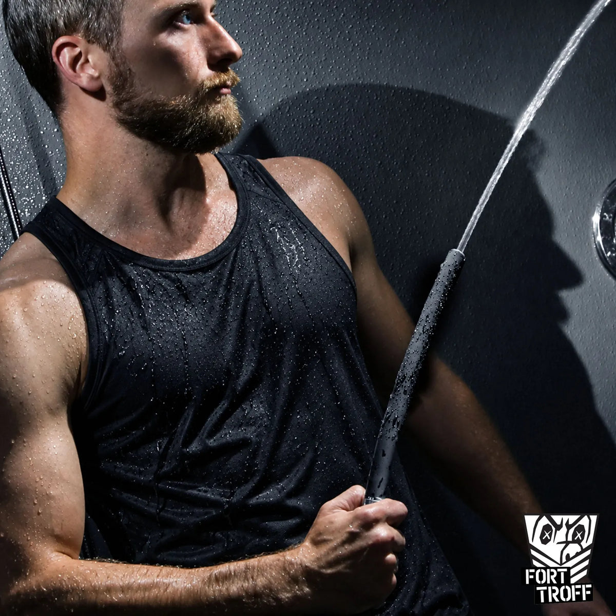 Bearded model in a soaked black tank top holding the Apex shower enema nozzle as water sprays, Fort Troff gay gear at Boyzshop