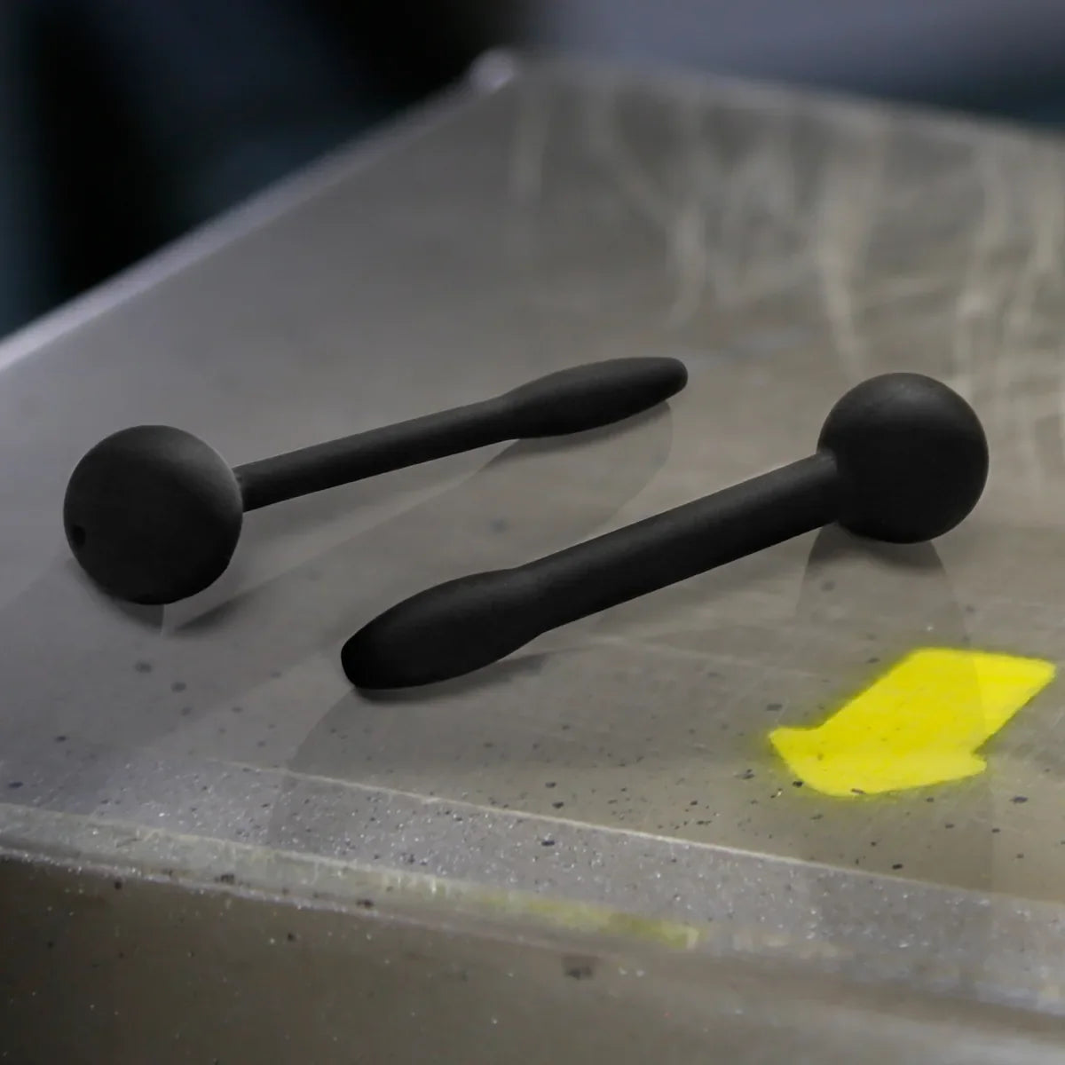 Pair of black penis plugs on a scuffed steel surface with a yellow arrow, Fort Troff gay gear at Boyzshop