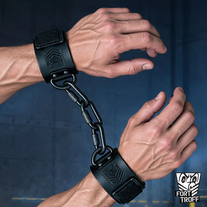 Leather wrist cuffs on both wrists linked by a black chain at the D-rings, dark blue backdrop, Fort Troff gay gear at Boyzshop