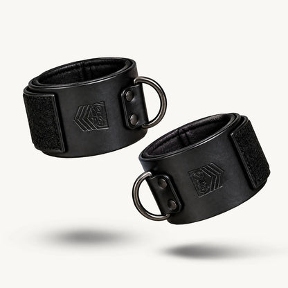 Pair of black leather wrist cuffs closed with Velcro, metal D-rings, white background, Fort Troff gay gear at Boyzshop