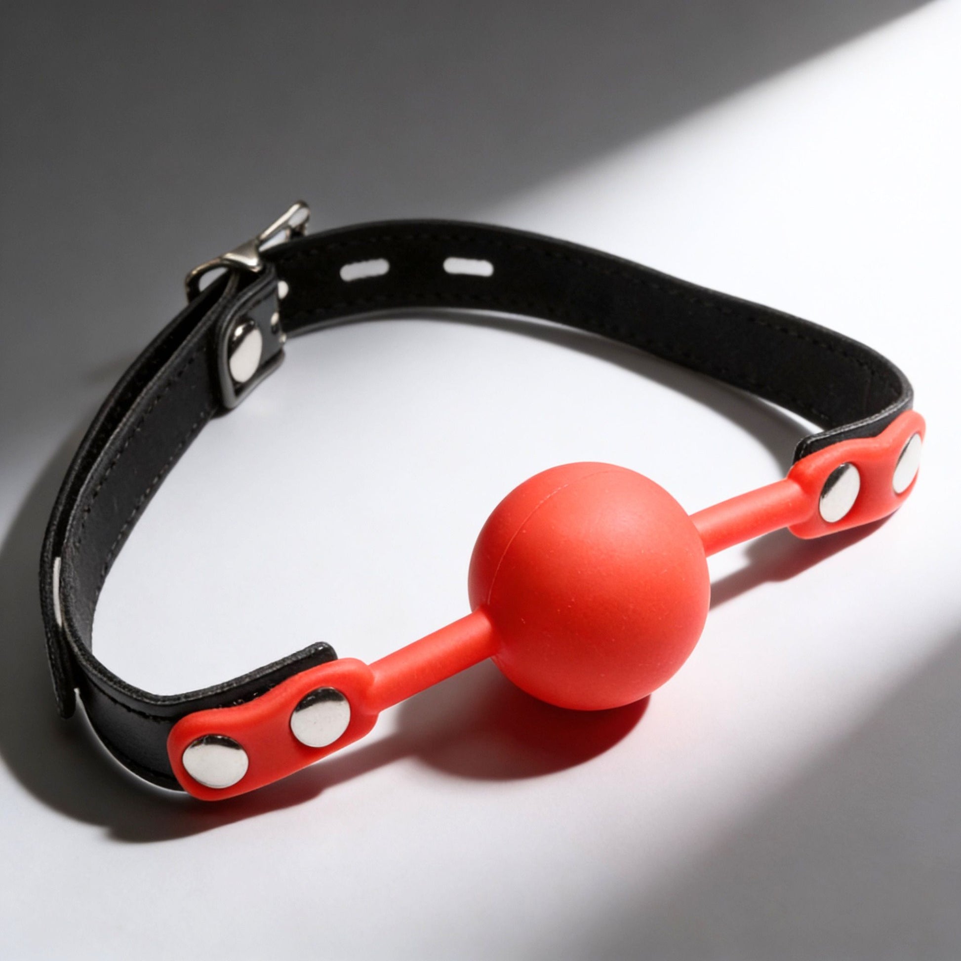 red silicone comfort ball gag with adjustable black strap and silver buckle hardware on gray background