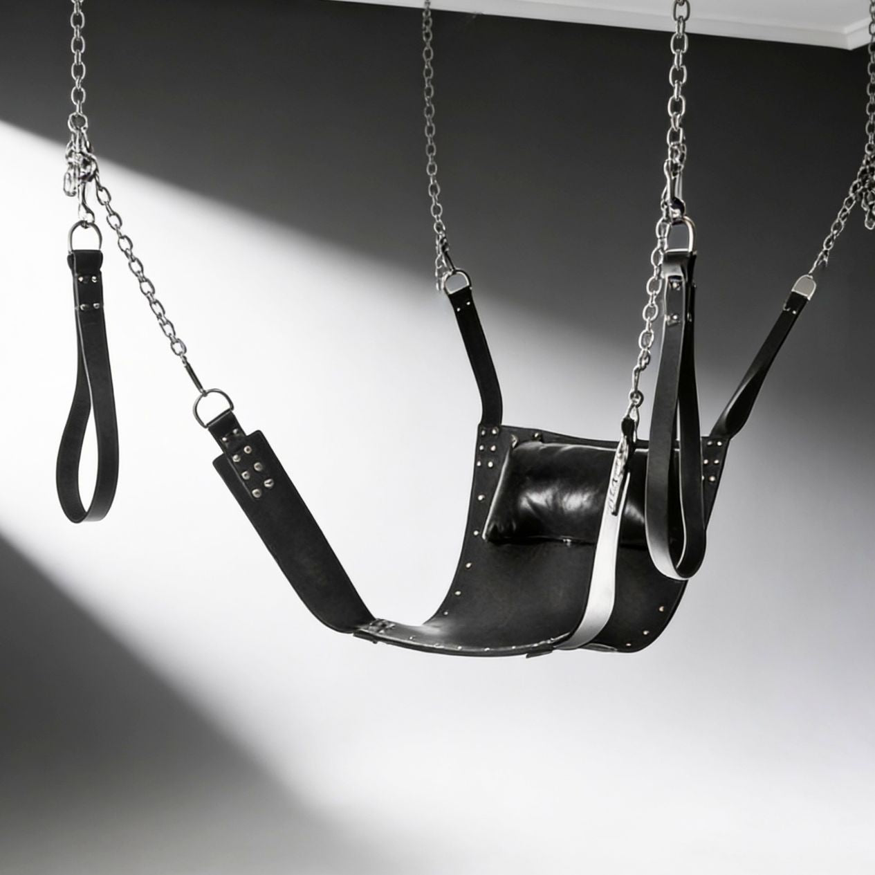 black leather bondage sling with stirrups, chains, and padded pillow suspended against gray background